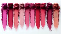 Smeared lipstick swatches in various shades of pink and red arranged in row on white background, showing texture and pigmentation of cosmetic products for makeup artists.