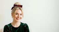 Young woman with blonde hair and fair complexion wearing dark green velvet top and burgundy sequined bow headband, smiling at camera against white background for portrait and lifestyle use.