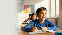 Happy young boy student smiling while writing in notebook at school desk with classmates in bright classroom for educational content.