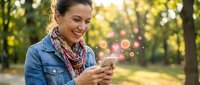 Happy middle-aged woman using smartphone in sunny park with bokeh lights, smiling while texting outdoors in casual denim jacket and scarf during golden hour.