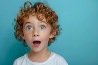 Young boy with curly ginger hair and bright blue eyes shows surprised expression with open mouth against turquoise background, wearing white t-shirt.