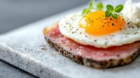 Open sandwich with dark rye bread topped with pink ham, sunny side up fried egg garnished with microgreens and black pepper on gray stone surface.