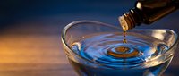 Essential oil dropping into glass bowl creating ripples against warm sunset background for aromatherapy wellness and natural healing treatments.