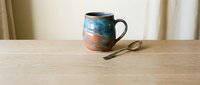 Handcrafted ceramic mug with blue and orange glazed finish sits on wooden table with vintage spoon, perfect for morning coffee or tea rituals.