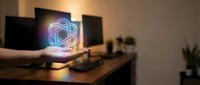 Hand holding glowing neon atom symbol above wooden desk with computer monitors, representing science, technology, innovation, and research in modern workspace environment.