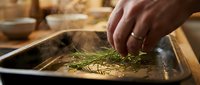Chef's hand sprinkling fresh rosemary herbs into a dark baking pan during food preparation in a professional kitchen setting.
