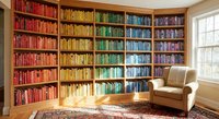Cozy home library with rainbow organized bookshelves featuring colorful book spines arranged by color from red to purple with comfortable reading chair.