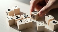 Hand placing wooden cube with brain icon among scattered blocks featuring various symbols including question marks, lightbulb, and communication icons on desk.