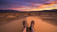 Hiker's feet in boots resting on golden sand dunes at sunset, with dramatic purple and orange sky over distant mountains, adventure travel perspective.