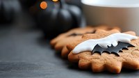 Homemade gingerbread cookies decorated with black and white bat shaped sugar icing on dark background with Halloween pumpkins and bokeh lights.