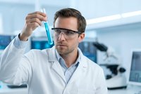 Male scientist in white lab coat examining blue liquid in test tube wearing safety glasses in modern laboratory for research and development.
