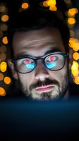 Caucasian male developer with glasses reflecting digital screen glow against bokeh lights background, close up portrait showing screen reflection in eyeglasses.