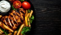 Grilled chicken breast with crispy french fries, fresh tomatoes, and creamy sauce on dark wooden background. Restaurant food styling for menu design.