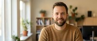Smiling middle-aged Caucasian man with beard in brown sweater sitting in modern home office interior with natural lighting for lifestyle content.