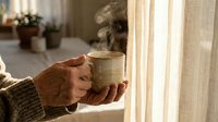 Senior woman hands holding steaming hot coffee mug by sunny window in cozy home interior morning light relaxation comfort lifestyle.