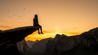 Silhouette of person sitting on rocky cliff edge during golden sunset with birds flying overhead, mountain landscape background for adventure and freedom concepts.