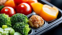 Fresh healthy meal prep container with broccoli florets, cherry tomatoes, yellow bell pepper and turkey meatball on black plastic tray, macro food photography.