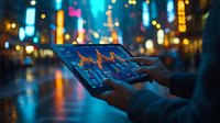 Hands holding digital tablet displaying financial charts and graphs against blurred urban night cityscape with colorful bokeh lights and reflections.
