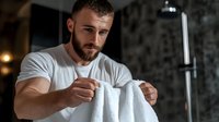 Young bearded Caucasian man in white t-shirt holding clean white towel in modern bathroom with dark tile walls, focused on personal hygiene and grooming.