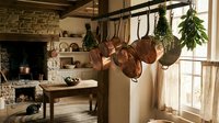 Rustic farmhouse kitchen with copper pots hanging from wooden beam, stone fireplace and herb bundles drying by sunny window for cozy home cooking.
