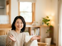 Middle-aged woman with dark hair and East Asian ethnicity wearing a beige knit sweater, smiling warmly while gesturing with open hands in a bright, modern living room with wooden furniture and plants.