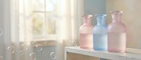 Pastel colored glass bottles in soft pink and blue tones on wooden surface with soap bubbles floating in bright sunlit bathroom interior.