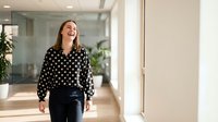Young woman with long brown hair and fair complexion wearing a black and white polka dot blouse and dark pants, walking through a modern office corridor with glass partitions and natural lighting.