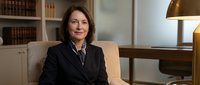 Professional middle-aged Caucasian businesswoman in dark suit sitting confidently in elegant office with bookshelf and warm lighting for corporate presentations.