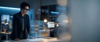 Young Asian male professional wearing glasses and dark blazer working with holographic data visualization and digital interface in modern tech environment.