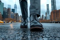 Black leather dress shoes on wet city street with urban skyline and skyscrapers in background during evening. Low angle perspective captures reflection on rainy pavement.