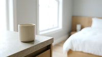 Ceramic mug with hot beverage on wooden nightstand in bright bedroom with natural light from window and bed in background.