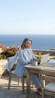 Middle-aged woman with light brown hair working on laptop at wooden table overlooking sea, wearing light blue blazer, surrounded by pink flowers and coastal scenery for remote work and lifestyle concepts.