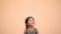 Young girl with dark hair and South Asian ethnicity, wearing a patterned top, looking upward with wonder and curiosity against a warm beige background. Suitable for child development, education, and aspirational concepts.