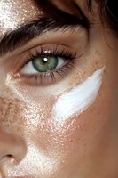 Close-up of green eye with freckles and white moisturizer cream streak on glowing skin, showing skincare and beauty treatment application.