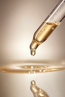 Glass dropper with golden liquid forming bubbles and droplets against beige background, reflection on surface creates artistic composition for cosmetic and wellness products.