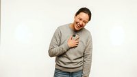 Man with dark hair and beard wearing glasses and beige sweatshirt, hand on chest, smiling warmly against white background for portrait and lifestyle use.
