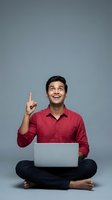 Young man of South Asian descent wearing red shirt sitting cross-legged with laptop, pointing upward with excited expression against gray background for business and technology concepts.