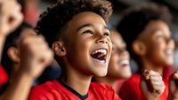 Young African American boy in red jersey cheering enthusiastically with raised fist at sports event, showing pure joy and excitement while watching game with crowd.