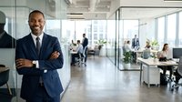 Confident male professional in navy business suit standing with crossed arms in modern office environment with colleagues working at desks behind glass partitions and windows.