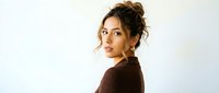 Young attractive mixed-race woman with curly hair in messy bun wearing brown ribbed sweater looking over shoulder against white background for fashion.