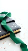 Black leather notebook with green satin ribbon and scattered confetti pieces on white background for celebration and party planning concepts.
