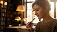 Young woman with curly hair writing in notebook at cozy cafe with warm lighting. Thoughtful female author journaling in comfortable indoor setting.