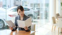 Young Asian businesswoman in gray suit reviewing documents at table in bright modern office or showroom.