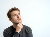 Thoughtful young man with brown hair and facial hair wearing dark gray sweater, hand on chin, looking upward in contemplative pose against white background for business and lifestyle content.