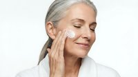 Mature Caucasian woman applying facial cream with closed eyes, enjoying skincare routine. Anti-aging beauty concept for cosmetic products and wellness.