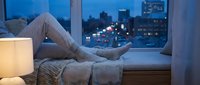 Woman relaxing in cozy pajamas by window with city lights view at evening. Comfortable home lifestyle and urban living concept for wellness content.
