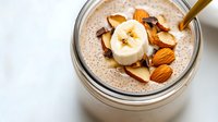 Creamy chia seed pudding topped with fresh banana slices, almonds and chocolate chips in glass jar with golden spoon for healthy breakfast or dessert.