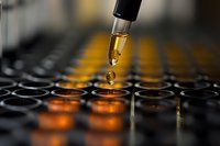 Droplet of amber liquid falling from pipette into laboratory microplate wells with orange reflective glow, macro photography showing scientific research process.