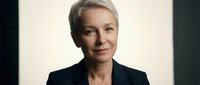 Professional mature Caucasian businesswoman with short blonde hair in dark suit jacket smiling confidently against neutral background for corporate use.