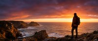Silhouetted hiker stands on rocky cliff overlooking dramatic ocean sunset with golden sky and crashing waves, adventure travel and outdoor exploration concept.